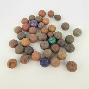 May include: A collection of numerous spherical marbles in various earthy tones including shades of brown, tan, gray, and muted orange, with a few individual marbles featuring speckled blue, green, or purple textures scattered across the surface.