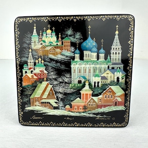 May include: A rectangular black lacquer box featuring a detailed hand-painted scene of a Russian town with blue-domed churches, wooden houses, and snowy landscapes. The border is decorated with gold filigree patterns and Cyrillic text at the base.