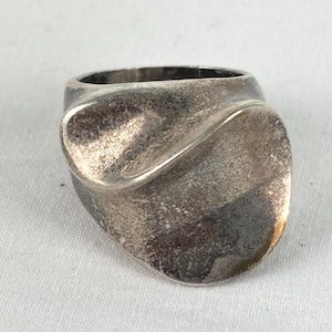 May include: A silver-toned ring with a wide, asymmetrical band featuring a smooth, curved, and folded surface design. The metal has a slightly weathered, matte finish with subtle variations in texture across the rounded, organic shape.