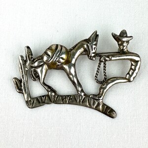 Puede incluir: Broche de metal plateado que representa una figura de vaquero sosteniendo una cuerda atada a un burro. El burro está de pie sobre una rama junto a un cactus. El broche tiene un diseño vintage con temática del Oeste.