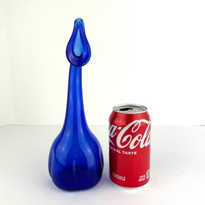 May include: A tall, cobalt blue glass decanter with a long, slender neck and a flared, teardrop-shaped opening. Next to it stands a red 12 fluid ounce Coca-Cola aluminum can, which serves as a scale reference for the height of the glass vessel.