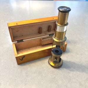 May include: A vintage brass microscope and wooden storage box. The microscope has a cylindrical body with a lens at the top. The wooden box is open, revealing its interior. The box is made of light-coloured wood.