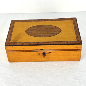 May include: A rectangular wooden box featuring a light yellow finish with a dark brown oval inlay on the lid. The lid is bordered by a thin, intricate geometric pattern. The front panel includes a small, diamond-shaped keyhole escutcheon.