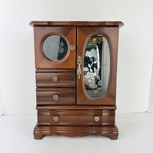 May include: A wooden jewelry armoire with a dark brown finish. It features a round glass window with a floral design, an oval mirror with a floral etching, and multiple drawers with heart-shaped knobs. The armoire has a decorative handle and a curved base.