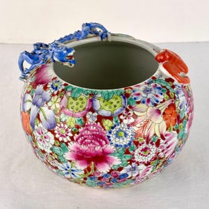 May include: A round ceramic bowl featuring a vibrant floral pattern in shades of pink, purple, blue, and green on a red background. The rim is adorned with a sculpted blue dragon on one side and a sculpted orange creature on the opposite side.
