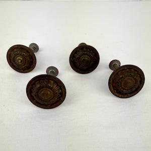 May include: Four antique bronze cabinet knobs with a detailed floral design. Each knob has a round, ornate face and a small, silver-colored stem for mounting. The knobs are arranged on a white surface.