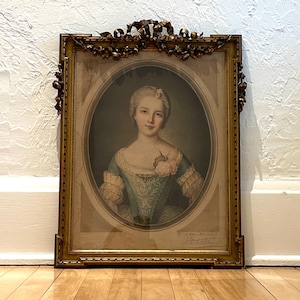 May include: An antique framed portrait of a woman in a blue dress with a pink flower. The portrait is in an oval frame, surrounded by a gold ornate frame with decorative elements. The artwork is displayed on a wooden floor.