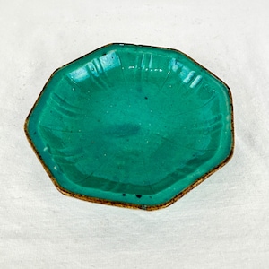 Puede incluir: Un plato de cerámica octogonal con un esmalte turquesa vibrante y un acabado craquelado sutil. Los bordes del plato están ligeramente elevados y definidos por hendiduras verticales, mientras que el borde exterior muestra un tono marrón.