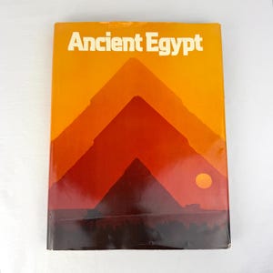 May include: A rectangular book titled Ancient Egypt with a cover featuring a gradient illustration of pyramids in shades of yellow, orange, and dark red, with a small yellow sun circle on the right side against a dark brown horizon.