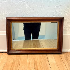May include: Rectangular mirror with a dark brown wooden frame and a gold inner trim. The mirror reflects a person wearing black pants. The mirror is set against a white wall and rests on a wooden floor.