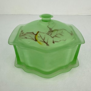 Könnte beinhalten: Hellgrüne, vintage Schmuckschatulle aus Glas mit Deckel. Die Box hat eine quadratische Form mit einem gewellten Rand und einem kleinen Griff auf dem Deckel. Der Deckel ist mit einem bemalten Blatt- und Beerendesign verziert.