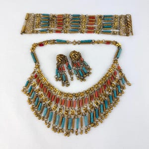 Pode incluir: Um conjunto de joias composto por uma pulseira larga, um colar de várias camadas e dois brincos pendentes. Cada peça é feita de metal dourado com contas cilíndricas alternadas nas cores turquesa e coral e pequenas esferas metálicas pendentes.