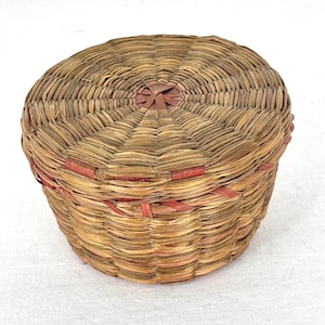 May include: A round, woven basket with a lid. The basket is made of natural-coloured reeds with red accents around the rim and on the lid's centre. The basket is approximately 13 cm in diameter and 8 cm tall.
