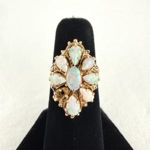May include: A gold ring featuring a central oval opal surrounded by eight pear-shaped opals arranged in a floral pattern. The metal setting has an intricate, openwork design that frames each gemstone, creating a cluster of iridescent stones.