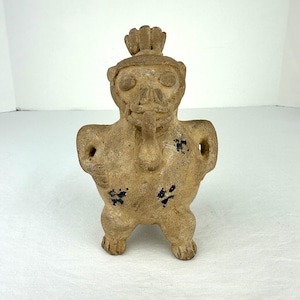 May include: A tan-colored clay figurine depicting a stylized creature with a prominent headdress, large circular eyes, and a protruding tongue. The figure has a rounded torso, small arms with visible openings, and black paint spots on the body.