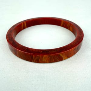 May include: A circular bangle bracelet featuring a polished, smooth surface with a marbled pattern of deep red, burnt orange, and amber tones. The bracelet has a thick, solid band construction with a glossy finish throughout the entire piece.