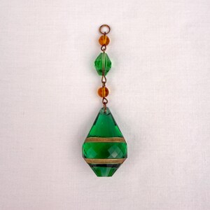 May include: A green glass pendant with a teardrop shape, featuring horizontal gold bands and faceted surfaces. It is strung with amber-colored beads and a small metal ring at the top, creating a decorative hanging ornament.