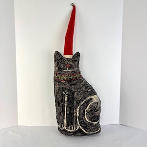 May include: A decorative cat-shaped pillow with a red hat and a bell on top. The cat is primarily dark gray with white and tan accents. It has a festive collar with red berries and green leaves. The pillow is designed to sit upright.