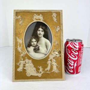May include: A rectangular gold-toned picture frame featuring an oval center cutout containing a black and white portrait of two individuals. The frame is decorated with embossed figures of cherubs and people. A red Coca-Cola can is placed beside it.