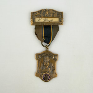 Puede incluir: Una insignia de metal color bronce del decimoquinto congreso nacional de la American Legion de 1933 en Chicago. Presenta una barra superior con un águila, una cinta con una franja central azul y un colgante en forma de escudo con figura.