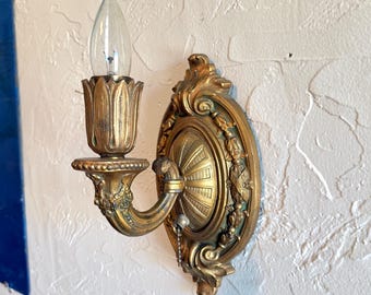 May include: A gold-toned wall sconce featuring an oval backplate with ornate scrollwork and a central fluted design. A curved arm supports a candle-shaped light fixture with a petal-like base and a clear glass bulb. A small pull chain hangs below.