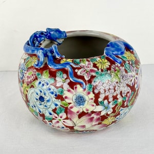 May include: A round ceramic bowl featuring a deep red background decorated with a colorful floral pattern of pink, blue, yellow, and white blossoms. A sculpted blue dragon figure is wrapped around the upper rim of the bowl.