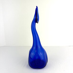 May include: A tall, vibrant cobalt blue glass sculpture featuring a rounded, bulbous base that tapers into a long, slender, curved neck. The top of the neck terminates in a delicate, leaf-like or petal-shaped finial, creating a fluid, organic form.