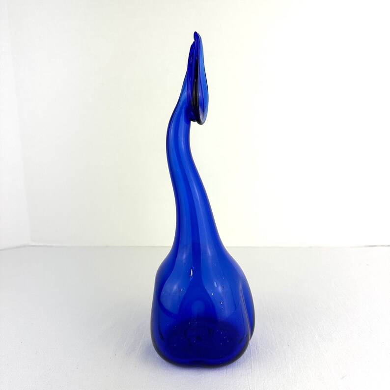 May include: A tall, vibrant cobalt blue glass sculpture featuring a rounded, bulbous base that tapers into a long, slender, curved neck. The top of the neck terminates in a delicate, leaf-like or petal-shaped finial, creating a fluid, organic form.