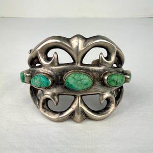 May include: A wide silver cuff bracelet featuring an intricate openwork design with curved metal elements. The centre of the bracelet is adorned with three oval-shaped, green turquoise stones with dark veining set horizontally in metal.