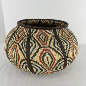 May include: A round woven basket with a dark brown rim and vertical stripes. The basket features a repeating pattern of cream, red, and blue diamond shapes. The basket is likely used for storage or decoration.