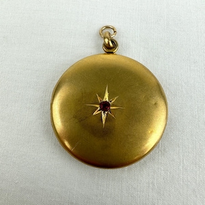 May include: A circular, gold-coloured locket featuring a starburst design with a small, red gemstone at its centre. It has a loop at the top for a chain. The locket is a vintage item.