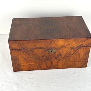 May include: A rectangular wooden box featuring a rich brown wood grain pattern with swirling dark brown markings. The box has a hinged lid and a central brass-colored metal keyhole escutcheon plate mounted on the front face of the lower section.