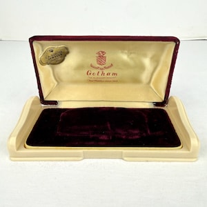May include: Open vintage jewelry box with a burgundy velvet interior and a cream-colored exterior. The box features the text "Gotham" and "J.A. Boelter Jeweler" on the inside. The box is open, revealing the velvet lining.