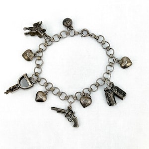 Op de afbeelding: Een zilveren bedelarmband met diverse bedels, waaronder harten, een zadel, een revolver en cowboylaarzen. De armband is gemaakt van aan elkaar gekoppelde cirkels en heeft een sluiting. De bedels zijn gedetailleerd en hebben een antieke uitstraling.