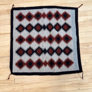 May include: Square woven textile with a repeating diamond pattern in red, navy, and gray. The diamonds are arranged in horizontal rows against a light gray background, framed by a dark border. Small tassels are visible at each corner.