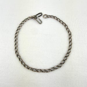 May include: A silver-toned chain with an oval link design. The chain is open, with a clasp on one end. The chain is likely made of metal and is displayed on a white surface. The chain is a jewelry item.