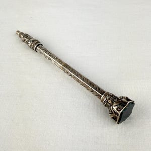 May include: An antique silver pencil with detailed floral and leaf patterns. The pencil features a dark green, hexagonal stone at the base and a textured tip. The pencil is approximately 15 cm long.