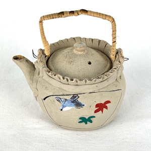 May include: A beige ceramic teapot with a woven handle. The teapot features a lid and spout, and is decorated with a blue bird and red and green leaf designs. The handle is made of woven straw.
