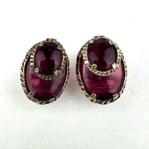 Op de afbeelding: Een paar vintage clip-on oorbellen. Elke oorbel heeft een grote, ovale, dieprode glazen steen gezet in een goudkleurig metalen frame. Het frame heeft een getextureerd, touwachtig ontwerp rond de randen, wat een vleugje vintage elegantie toevoegt.