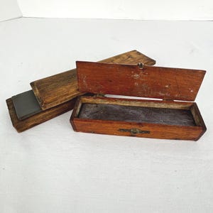 Peut inclure: Une boîte en bois rectangulaire ouverte avec un couvercle à charnière, révélant un intérieur sombre. Une boîte en bois plus petite séparée avec une pierre gris foncé est à côté. Le bois a une teinte brun-rougeâtre chaude.