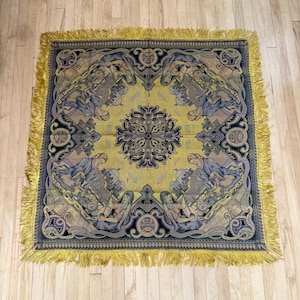 May include: A square woven tapestry featuring a central dark blue and black floral medallion on a mustard yellow background. The border displays repeating illustrations of figures in blue garments playing stringed instruments, finished with yellow fringe.