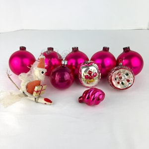 May include: A collection of vintage Christmas ornaments in shades of pink and silver. The ornaments include round, swirl, and decorative designs. A small, whimsical figure with a gift is also present.