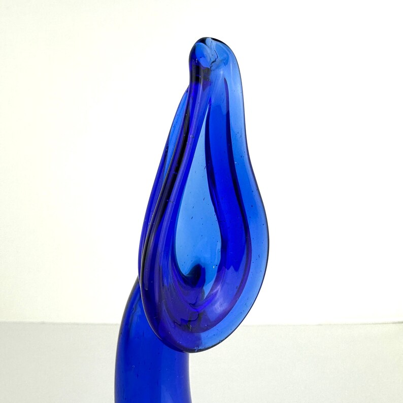 May include: A vibrant cobalt blue glass sculpture featuring a tall, slender base that curves upward into a wide, organic, teardrop-shaped opening. The translucent glass displays smooth, flowing lines and a glossy finish throughout the entire piece.