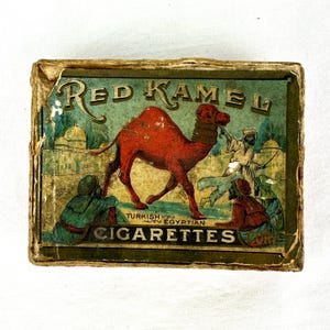 May include: A vintage Red Kamel Turkish Egyptian Cigarettes box. The box features a red camel, figures in traditional clothing, and the text "Red Kamel Cigarettes". The background is a faded green with architectural elements.