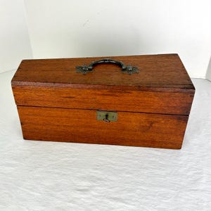May include: A rectangular wooden box with a dark brown finish. It features a metal handle and a small lock with a keyhole on the front. The box is designed with a classic, vintage aesthetic.