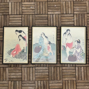 Może przedstawiać: Zestaw trzech oprawionych japońskich drzeworytów. Każdy z nich przedstawia kobiety w tradycyjnych strojach w pobliżu wody. Grafiki mają czarne ramy ze złotymi wewnętrznymi krawędziami. Grafika wykorzystuje stonowaną paletę barw niebieskiego, czerwonego i zielonego.