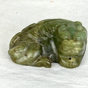 Può includere: Una statuetta di rana in giada scolpita in tonalità di verde. La rana è in posizione di riposo, con dettagli come la pelle strutturata e gli arti definiti. La statuetta è su una superficie bianca.