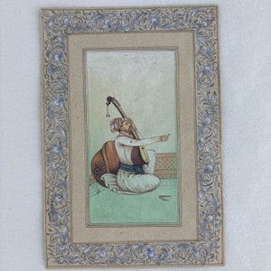 Puede incluir: Una pintura rectangular sobre papel que muestra a una persona con turbante y ropa blanca tocando un instrumento de cuerda mientras está sentada en un suelo verde. La obra está rodeada por un borde decorativo con motivos florales azules y grises.