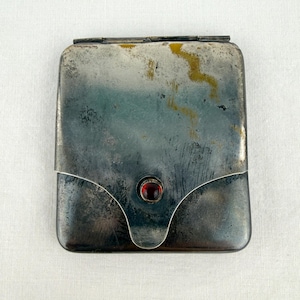 May include: A vintage silver-toned compact with a rectangular shape and a hinged lid. The compact features a decorative flap with a red gemstone accent. The metal has a tarnished appearance, adding to its antique charm.