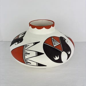 May include: A white ceramic vase with a red scalloped rim and decorative black and red geometric designs. The vase features stylized bird and abstract shapes. The vase is a unique piece of pottery.